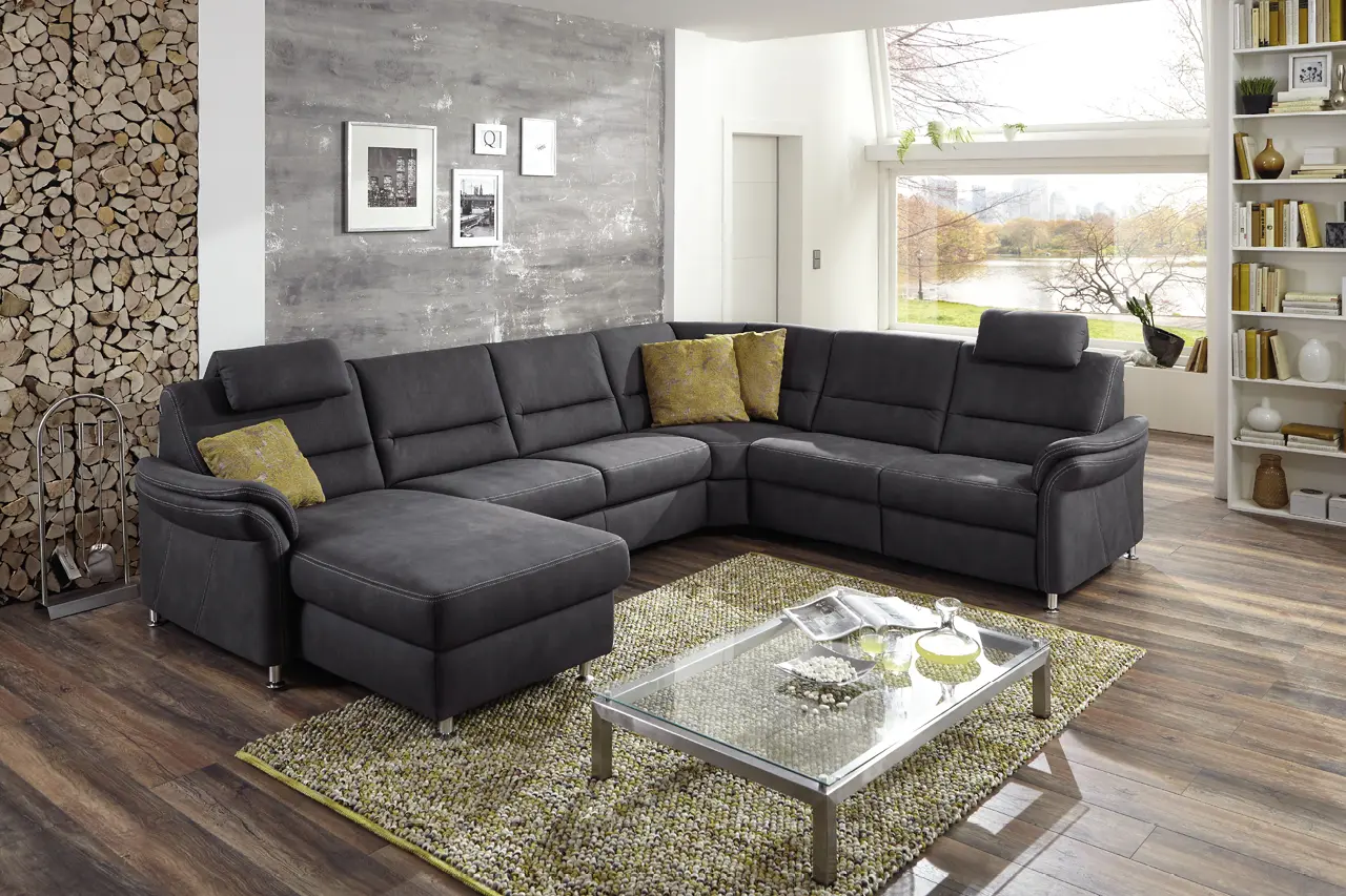 Dunkelgraue Wohnlandschaft mit Ottomane in modernem Wohnzimmer mit Holzfußboden, Teppich, Kaminholz und großem Fenster mit Blick auf Landschaft
